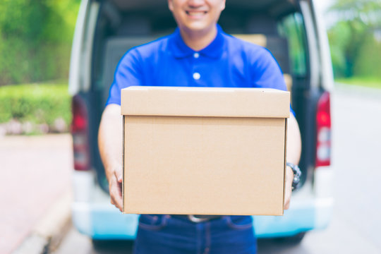 Delivery concept - Smiling happy young asian handsome male postal delivery courier man in front of cargo van delivering package holding box with service mind and blue uniform