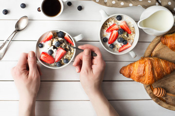Young woman holding muesli bowl in hands on white wooden background. Girl eating cereals breakfast with pumpkin seeds, oats,yogurt,strawberry and blueberry in bowl. Eye bird view