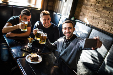 Handsome friends are drinking beer, doing selfie and smiling while resting in pub. Beer time.