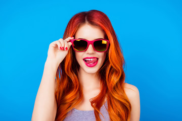 Funny carefree girl with ginger hair holding sunglasses and licking pink lips while standing on blue background