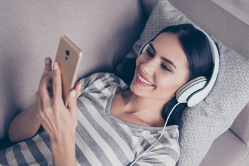 happy young cute pretty woman listen to music on headphones looking at smartphone while lying at home on couch