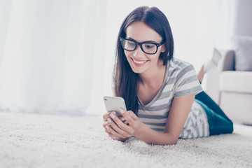 Young brunette pretty brunette girl in spectacles checking email and sms on smartphone while lying on the floor  at home