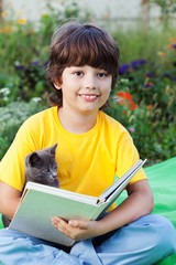 Boy reading book with kitten in the yard, child with pet reading magazine on the grass in park