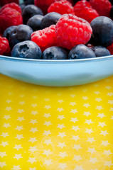 various kinds of fruit - berries raspberries, blueberries - in a blue bowl over yellow background with copy space 
