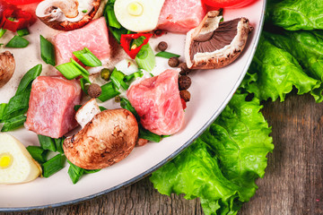 Raw meat in dish and frying pan, spices, knife on wooden table, vegetables, top view