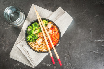 Healthy eating, diet. Buddha bowl hot salad in Asian style: noodles with soy sauce, spicy carrots, chicken, broccoli. On black concrete stone table, with chopsticks and water. Top view copy space