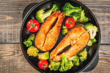 Healthy eating, diet. Baked grilled trout (salmon) with vegetable garnish - broccoli, tomatoes. In a portioned frying pan, on a wooden table. Top view copy space