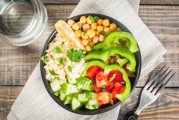 Healthy eating, diet. Vegetarian vegetable buddha bowl - couscous (quinoa), chickpeas, tomatoes, zucchini (cucumber), pepper, hummus, lemon and greens. On wooden table, with water, top view
