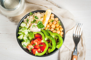 Healthy eating, diet. Vegetarian vegetable buddha bowl - couscous (quinoa), chickpeas, tomatoes, zucchini (cucumber), pepper, hummus, lemon, greens. On white wooden table, water, above copy space