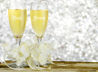 A pair of glasses filled with sparkling bubbly champagne and decorated with ivory colored satin ribbons and silk flowers on a wooden table in front of a sparkly, glittering silver bokeh background