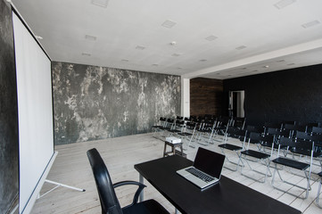 Room for lecture with a lot of dark chairs. Walls are white, loft interior. On the right there is a door. On the background there is a table with a laptop.