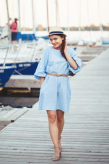 Portrait of white Caucasian brunette woman with tanned skin in blue dress by seashore lakeshore with yachts boats on background on water, lifestyle summer hobby leisure concept