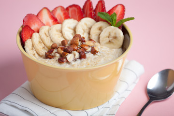 Bowl of oatmeal with a banana, strawberries, almonds, hazelnuts and butter on a pink background. Hot and a healthy dish for Breakfast, top view.