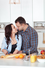 Couple making fresh organic juice in kitchen together. A young woman in a blue shirt slices a baguette. A man is hugging his girlfriend in a white kitchen. Romantic concept