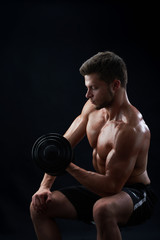 Fototapeta na wymiar Vertical studio shot of a handsome young shirtless athletic man with muscular body pumping iron exercising with heavy dumbbells on black background body torso biceps workout gym sport lifestyle.