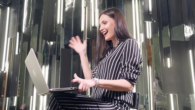 The girl businessman in a striped shirt in black and white sits behind a laptop and rejoices indicators on the screen.