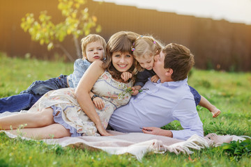 Naklejka na ściany i meble Family mom, dad, son and daughter are lying on a plaid on the green lawn. A boy hugs a woman, and a man gently kisses his daughter