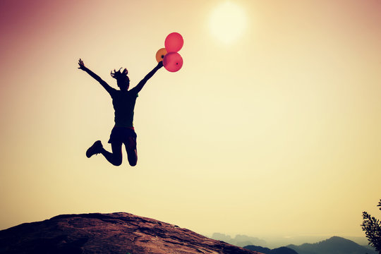 Young woman jumping with colorful balloons on sunrise mountain peak
