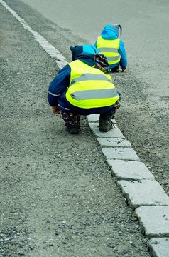 Two kids wearing reflective vests and playing on roadside.