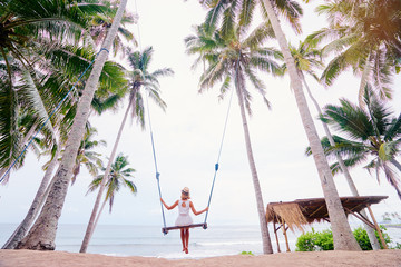 Vacation concept. Happy young woman in white dress and hat swinging at palm grove enjoying sea view.