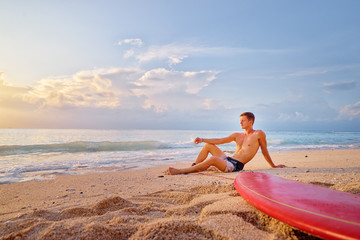 Surfing and vacation. Holiday on the beach. Relaxed young man sitting on the sand with surf board enjoying sunset.