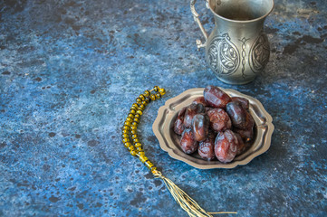 Dried date fruits or kurma served on a old vintage palte with ornaments and beads on a blue stone background, ramadan food concept.