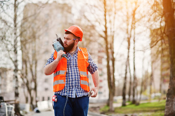 Portrait of brutal beard worker man suit construction worker in safety orange helmet against pavement with angular grinding machine in hand.