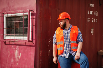 Brutal beard worker man suit construction worker in safety orange helmet sitting on red barrel.