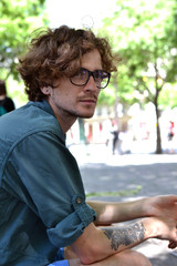 Beautiful young curly-haired guy in black-rimmed glasses travel in Europe on the background of the attractions in the Park, waiting for a date with a girl, resting, summer, spring