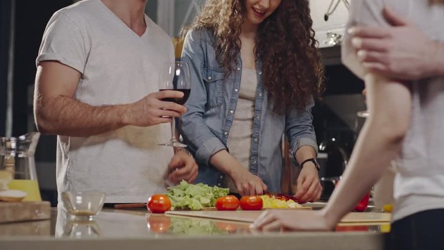 Tilt up shot of young man and woman having cooking party with friends in kitchen: girl chopping vegetables and man drinking wine and telling story