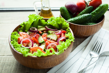 Rustic salad of fresh tomatoes, cucumbers, red onions and lettuce, dressed with olive oil and ground pepper in a wooden bowl. Top view