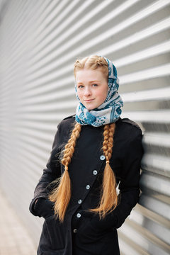 Young beautiful fashionable redhead woman with braids hairdo in blue white headcraft stylish denim black trench jacket posing outdoors against urban style background of metal strips