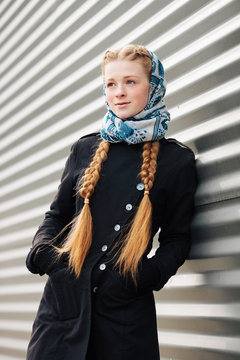 Young beautiful fashionable redhead woman with braids hairdo in blue white headcraft stylish denim black trench jacket posing outdoors against urban style background of metal strips