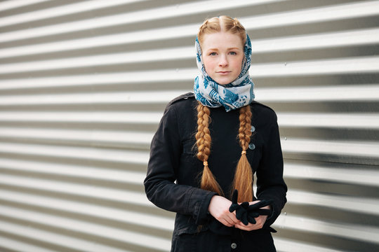 Young beautiful fashionable redhead woman with braids hairdo in blue white headcraft stylish denim black trench jacket posing outdoors against urban style background of metal strips