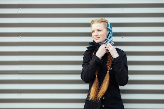 Young beautiful fashionable redhead woman with braids hairdo in blue white headcraft stylish denim black trench jacket posing outdoors against urban style background of metal strips