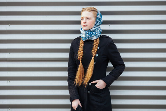 Young beautiful fashionable redhead woman with braids hairdo in blue white headcraft stylish denim black trench jacket posing outdoors against urban style background of metal strips