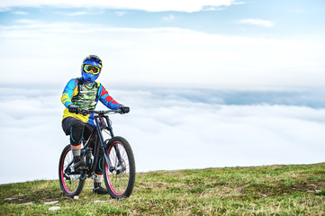 A young guy sits on a mountain bike atop a mountain, when below the mountains low clouds