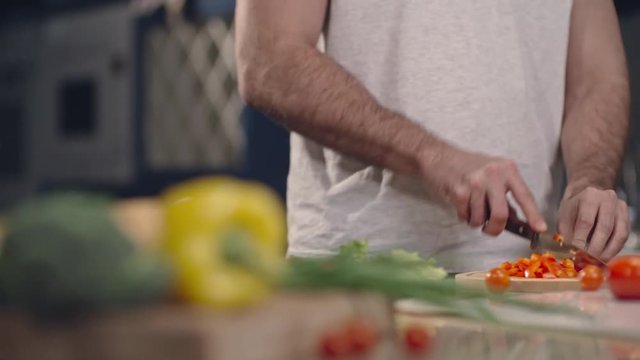 Tracking shot of man slicing bell pepper for salad on wooden cutting board on kitchen counter and putting it into glass bowl