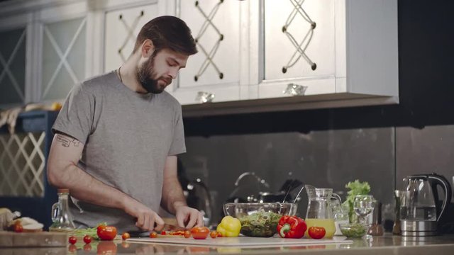 Tracking of bearded young man cutting cherry tomatoes and bell pepper and putting ingredients into glass bowl with salad