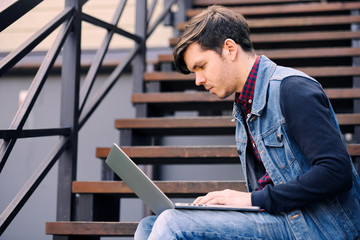 Young guy sits on the street on the stairs and uses a laptop. The concept of lifestyle and technology