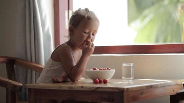 Little girl sits by the table and eating cherry tomato