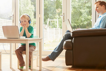 Boy at desk and father on sofa using laptops