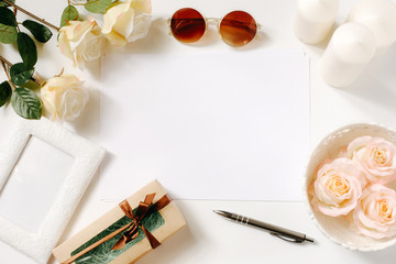 White desk with sunglasses, roses, candles, pen and vintage white tray. Empty sheet in the middle. Celebration card concept. Top view, flat lay, copyspace.