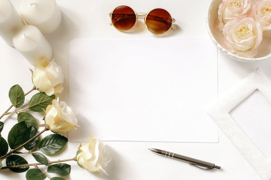 White desk with sunglasses, roses, candles, pen and vintage white tray. Empty sheet in the middle. Celebration card concept. Top view, flat lay, copyspace.