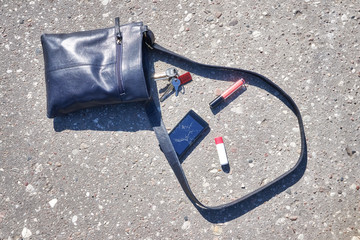 Handbag, cellular phone with broken screen, keys and lipstick on asphalt street, conceptual picture.