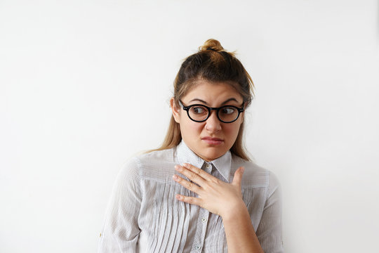 Headshot of disgusted female in striped shirt and glasses experiencing hostility to his colleagues in office and condemns her appearance. Hand on chest, raising brows. Negative people expressions