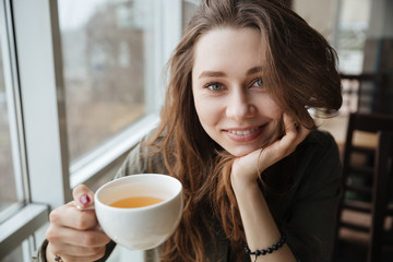 Cute happy young beautiful lady sitting in cafe drinking tea