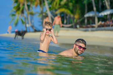 Happiness portrait in tropical water: seven years old blond boy sitting in water and his brown-haired bearded father is swimming like a crocodile. Both in sunglasses.