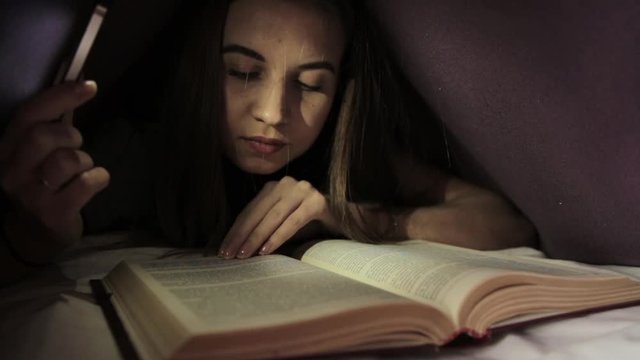 young girl using flashlight in dark for reading book under the blanket