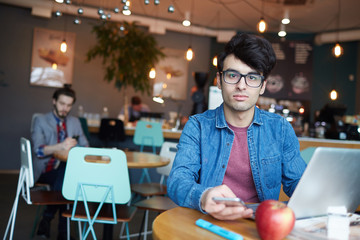 Fototapeta premium Portrait of handsome young man wearing glasses and jeans jacket looking at camera while working with laptop and smartphone at table in cafe